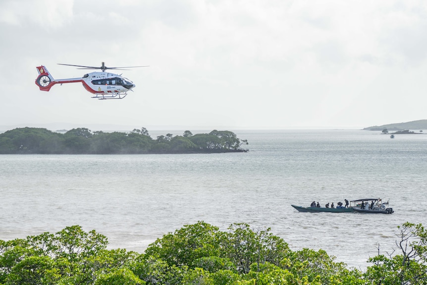 Several people in a small boat near a stretch of coastline. A helicopter flies above.