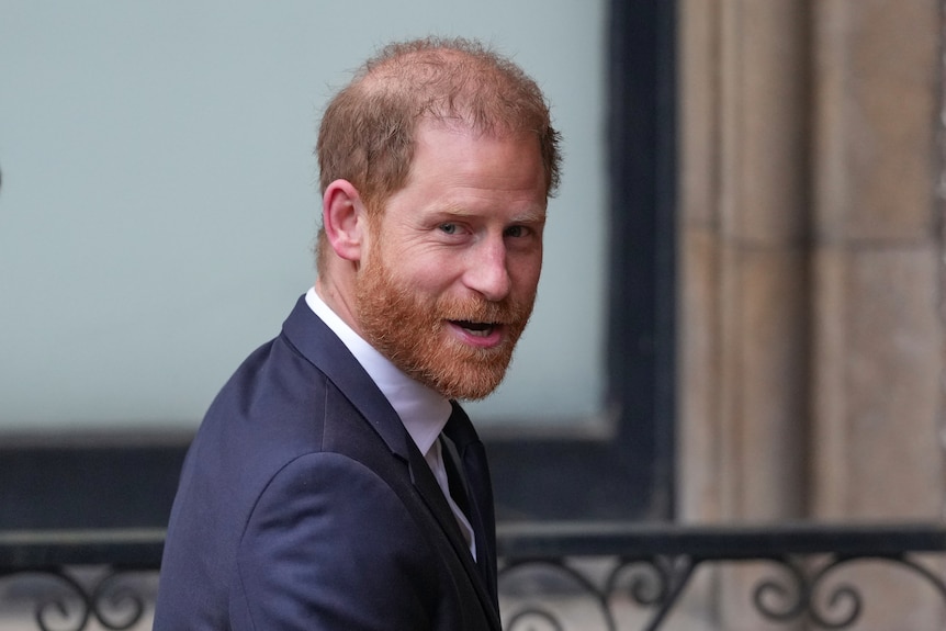 A close up of Prince Harry who looks over his shoulder as he speaks, while wearing a navy blazer