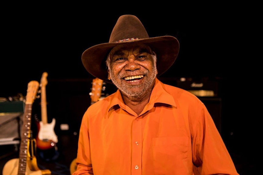 Warren H Williams wears an orange shirt, brown hat and he has a huge smile on his face. The are guitars in the studio behind.