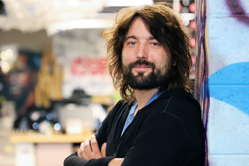 Tom, a man with long brown hair and a beard, smiles at the camera as he leans against a wall