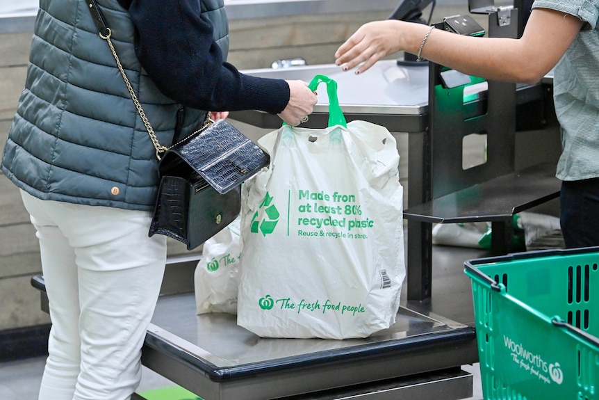 A person grabs a full Woolworths-branded reusable shopping bag from a supermarket check out-cashier. 