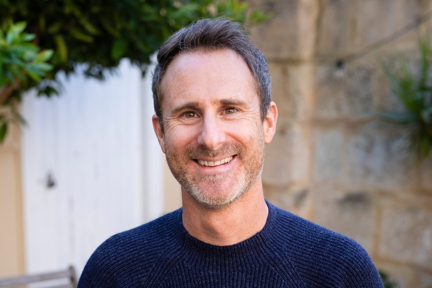 A portrait of Craig Silvey, 43, with short brown hair and stubble, smiling brightly with his hands in his pockets.