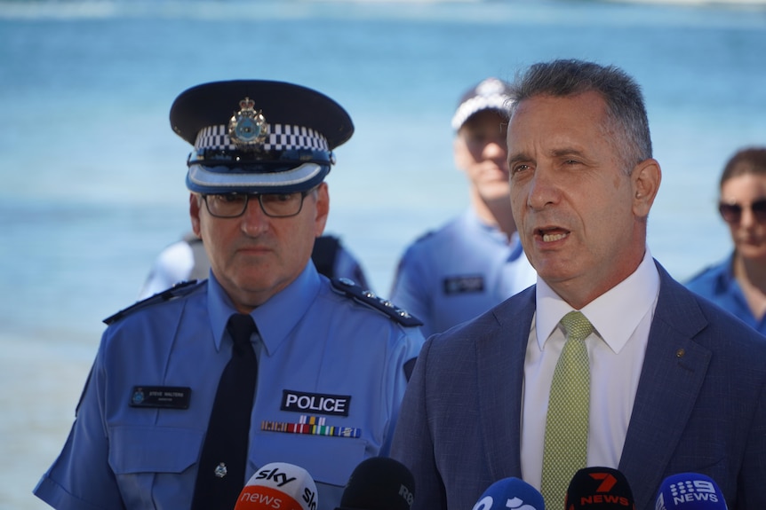 Paul Papalia stands in front of a police officer as he delivers a press conference with the ocean in the background