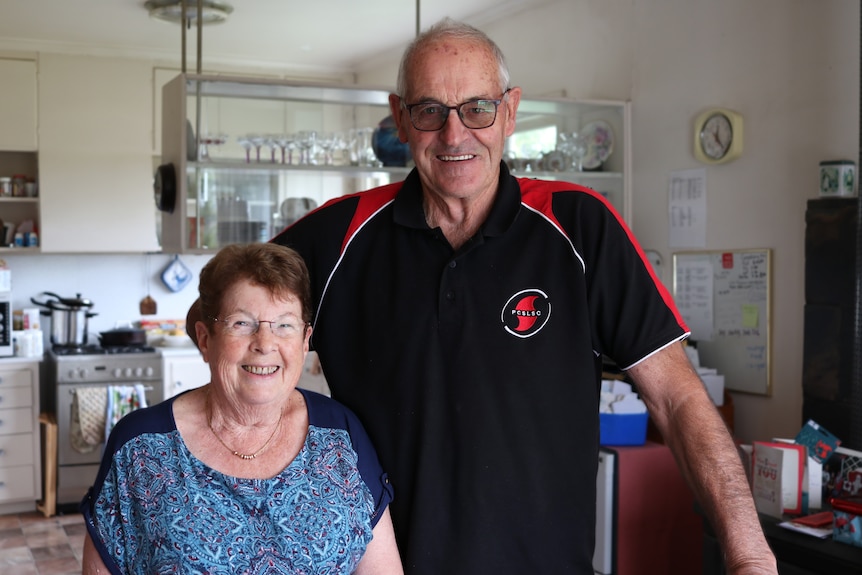A retiree aged couple smiling, older style kitchen behind them.