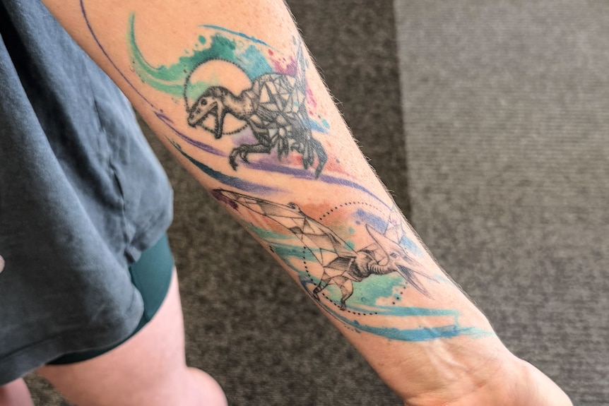 Woman holding our arm showing two colourful tattoos of dinosaurs
