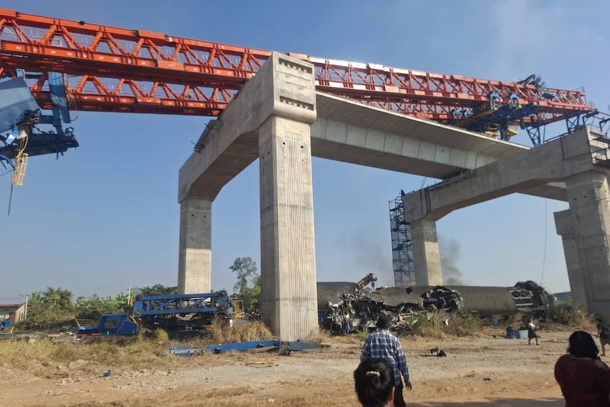 A large red crane has collapsed onto a concrete bridge. Underneath is a destroyed train carriage. People stand around. 
