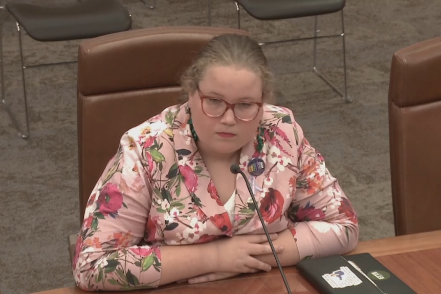 A woman in glasses and a floral shirt sits with her arms crossed at a wooden table..