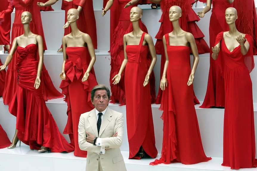 Valentino standing, arms folded, with a dozen mannequins behind him in red dresses.
