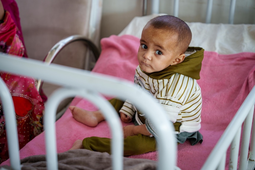 A young Afghan boy sits on a pink blanket in a bed in hospital.