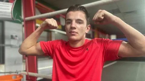 Simon Thake/BBC A young man with slicked back hair wearing a red sports t-shirt holds up his fists in a boxing pose