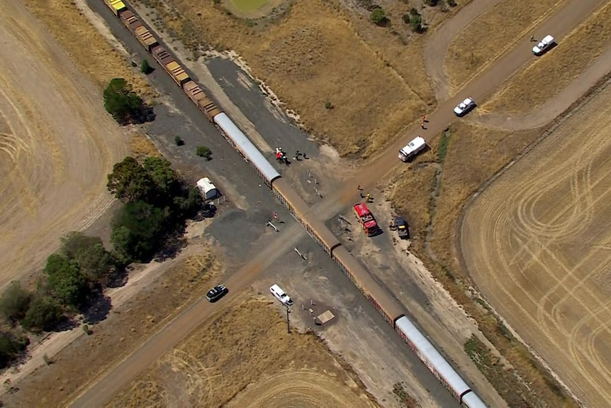 An aerial shot showing a long train stopped in the middle of a country intersection with a road, with emergency vehicles nearby.