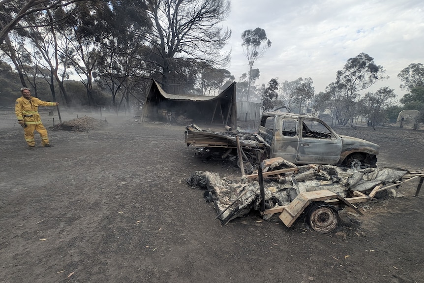 Firefighter in yellow uniform stands in front of the ruins of a metal shed looking at a burnt ute surrounded by blackened ground