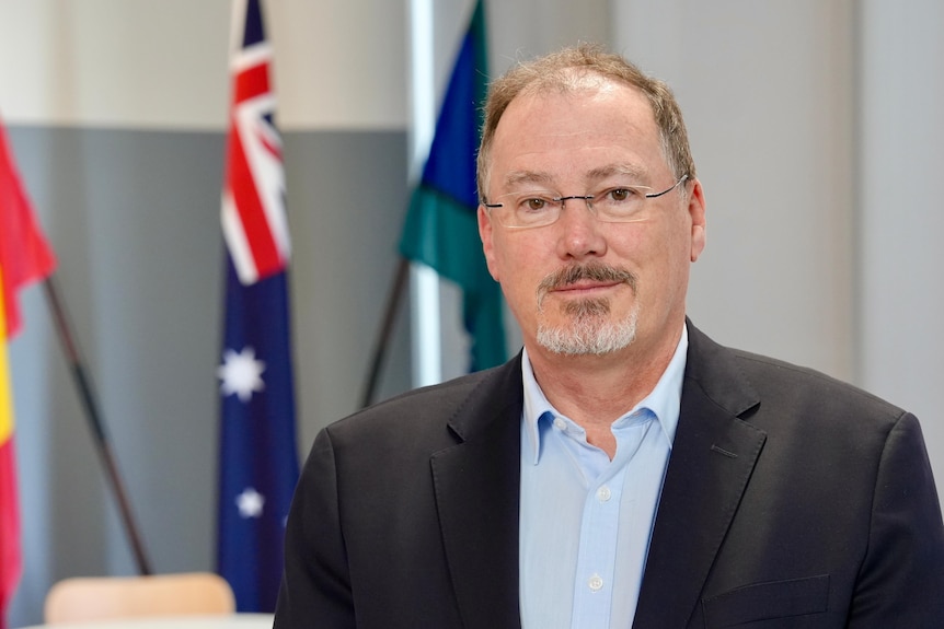 A man wearing a black blazer and blue shirt, wearing glasses, looks at the camera.