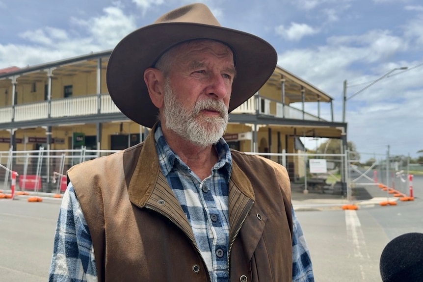 A photo of a man, with a gray beard, wearing a flannel shirt, brown vest and cowboy hand, standing in front of a pub