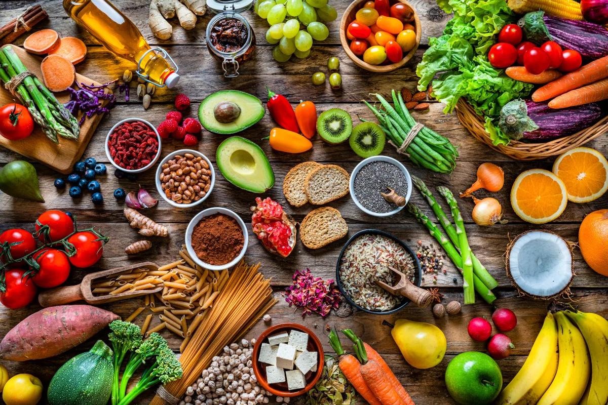 large group of multicolored fresh fruits, vegetables, cereals and spices shot from above on wooden background. The composition includes green apple, kiwi, pear, pomegranate, orange, coconut, banana, grape, berries, ginger, almonds, pistachio, olive oil, olives, goji berries, chia seeds, pinto beans, nutmeg, rosemary, radish, tomatoes, carrot, kale, avocado, onion, rice, cocoa powder, sweet potato, wholegrain pasta, tofu, lettuce, corn, broccoli, pepper, asparagus, green beans, among others.