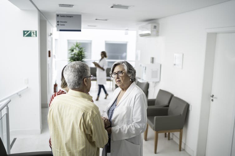 A senior man talks with a frustrated-looking health care provider in a hallway.