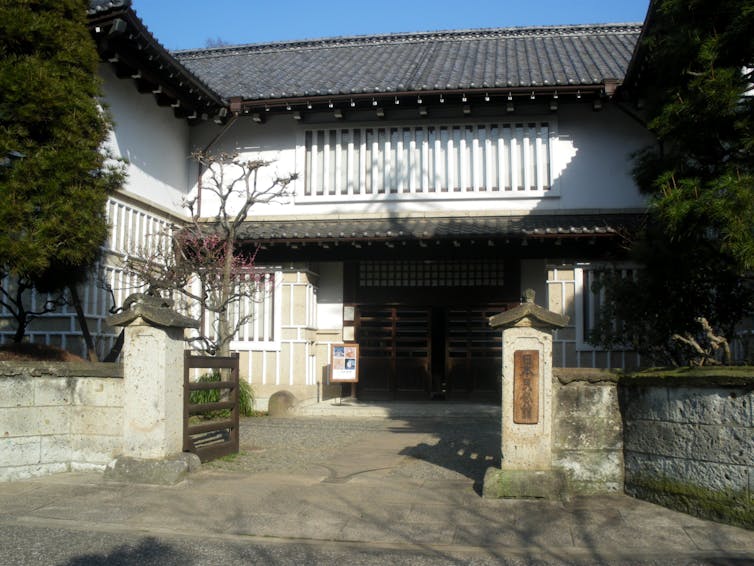 A view of the front of a traditional Japanese building with a dark roof and large wooden doors. There is a short stone fence in the foreground.
