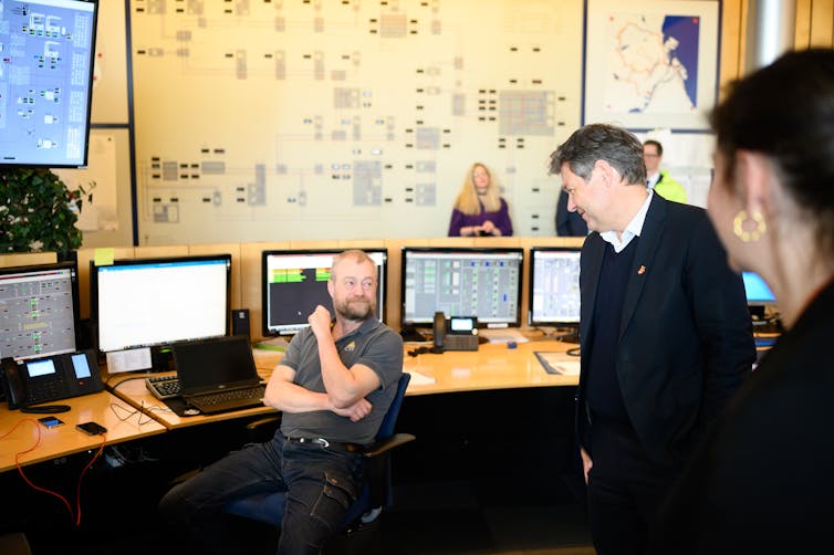 People talk in a room with many computer screens and large diagrams on the wall.