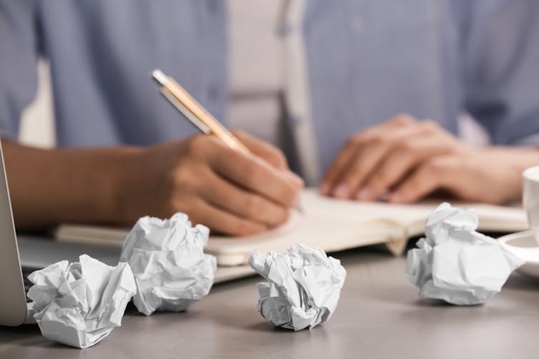 Close up of a person's hands writing with pen in a notebook, with crumpled up papers surrounding on the table