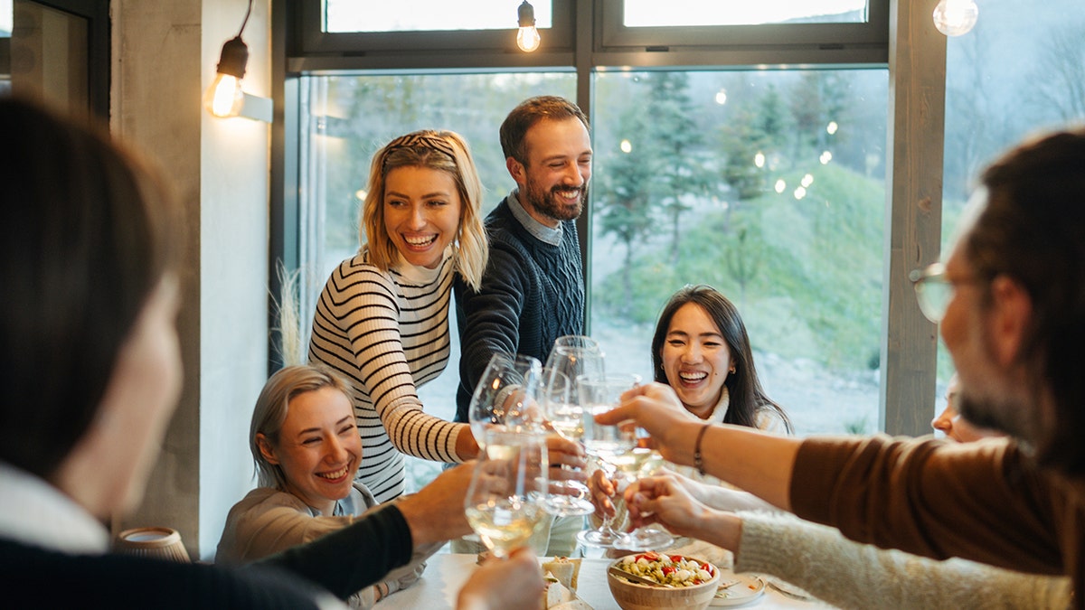 Group of friends smiling and clinking wine glasses around a dining table in a cozy, modern home setting.