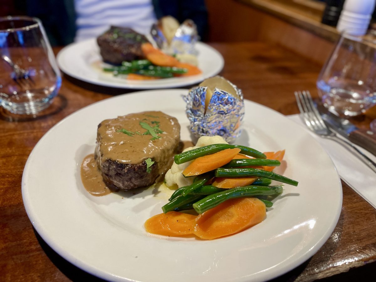 A white plate with eye fillet steak covered in gravy, a jacket potato in foil and steamed carrot, beans and cauliflower.