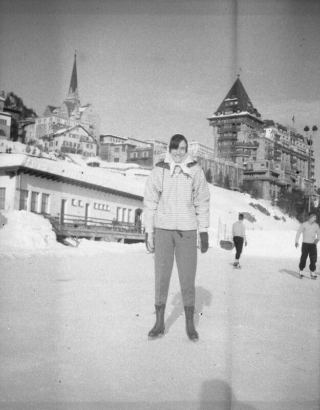 A person wearing winter clothes stands on an ice rink with snow-covered buildings, a church steeple, and a large tower in the background; others are skating nearby. The scene is bright, and the photo is in black and white.