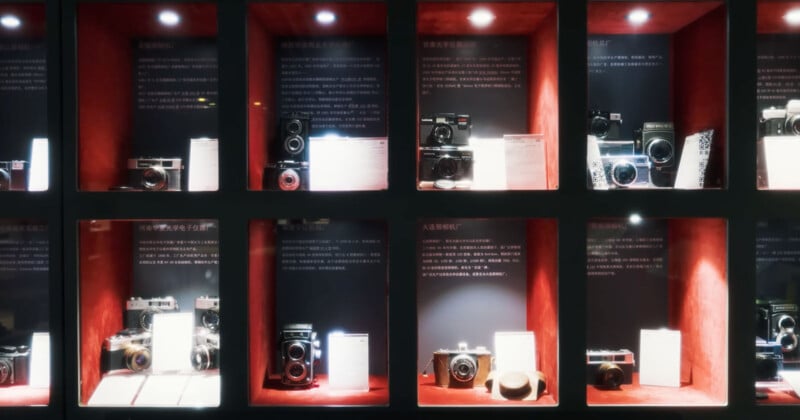 A display case with multiple compartments showcases various vintage cameras and accessories, each compartment illuminated by overhead lights, with informational plaques accompanying the items.