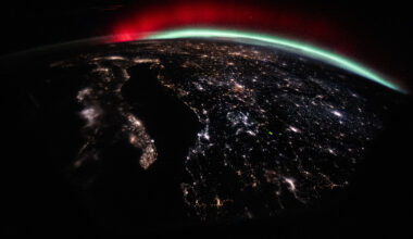 A green and red aurora streams across Earth’s horizon above the city lights of Europe.