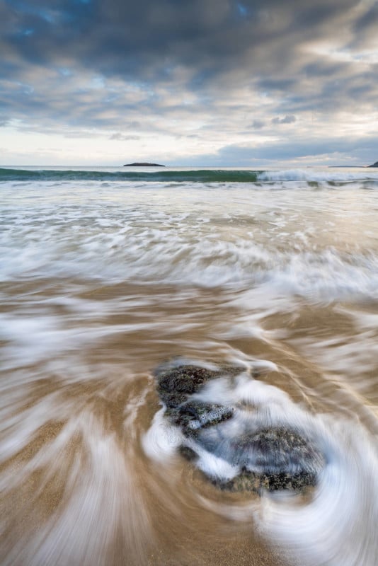 Waves swirl around rocks on a sandy beach under a cloudy sky, with distant land visible on the horizon. The water appears blurred, capturing the motion of the surf.