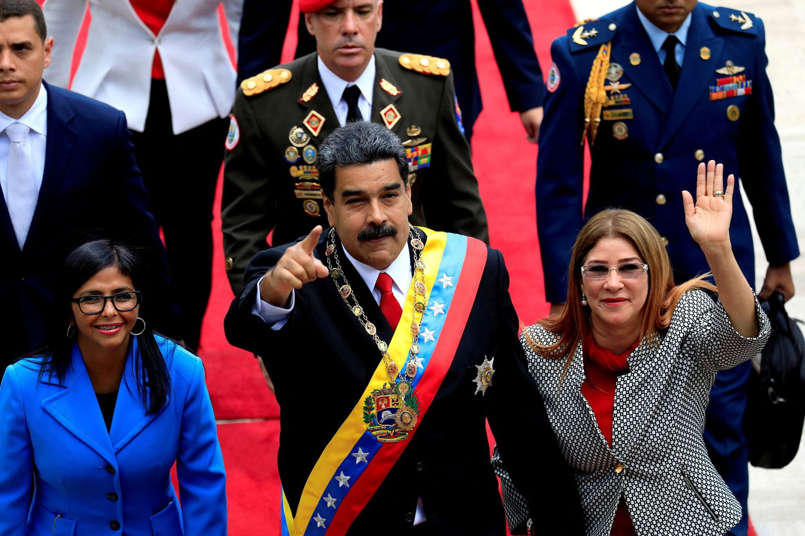A man in an ornate necklace and sash with Venezuelan colours points ahead. He holds the hand of a woman who waves. On his other side is a woman in a cobalt pea coat