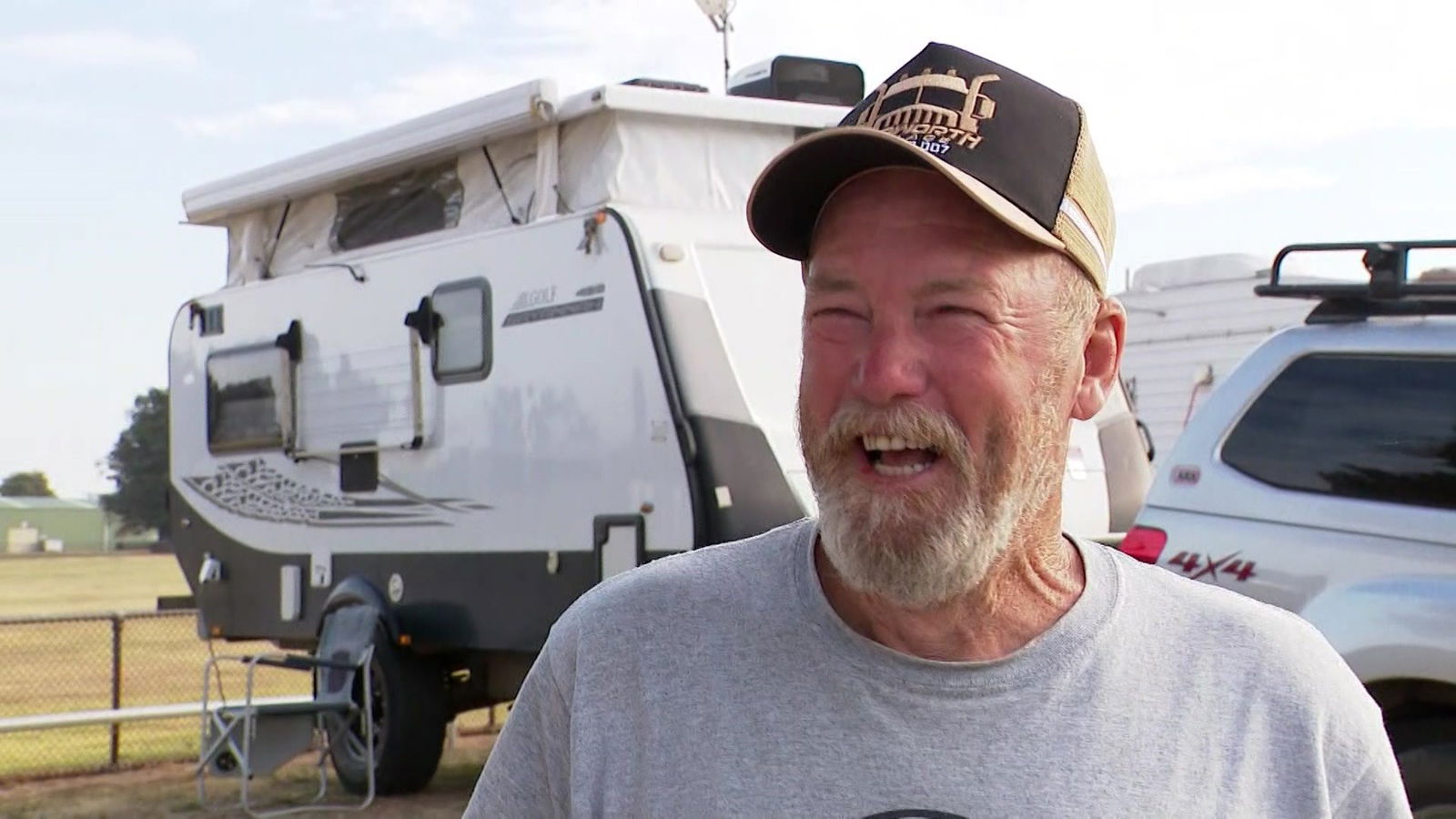 A man smiles outside a caravan, talking to a reporter.