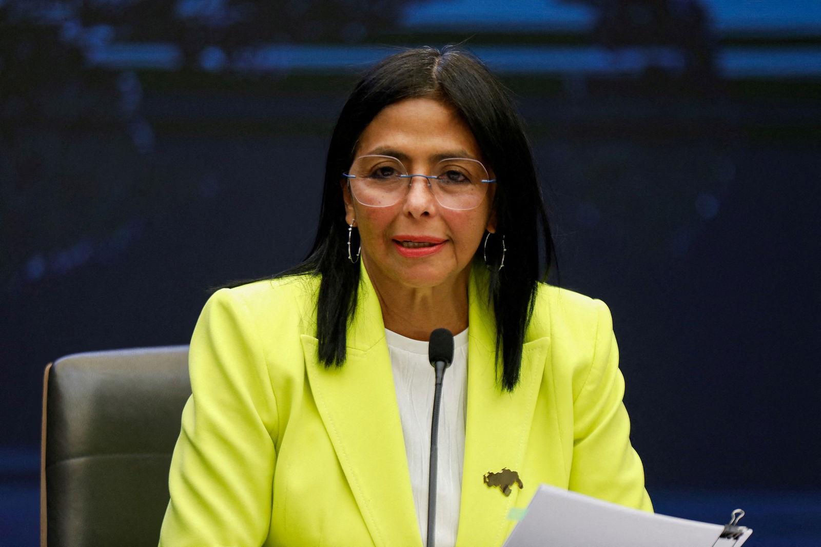 A woman with straight, black hair wears large-framed rimless glasses and a neon yellow blazer