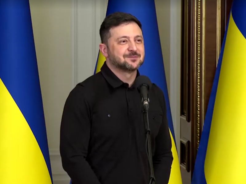 Zelenskyy stands in front of three Ukrainian flags with a small smile