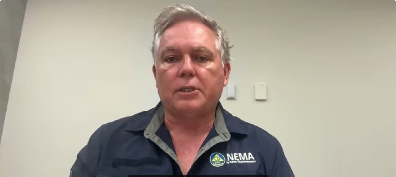 A man wearing a dark shirt labelled "NEMA" sits in front of a white wall as he speaks