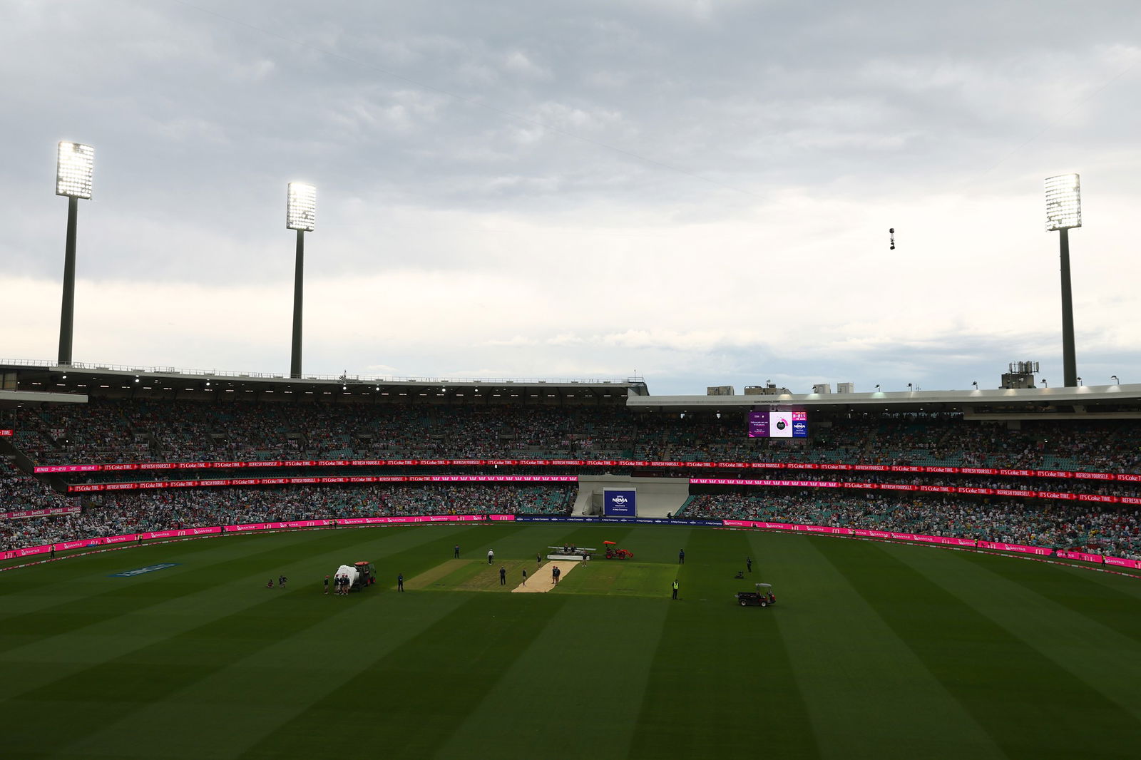 A landscape shot of a cricket field with grey clouds over it and floodlights on