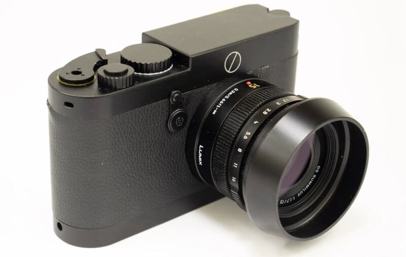 A black digital camera with a textured grip and a prominent lens, displayed on a white background. The lens has white markings and a red "15" indicating its focal length.