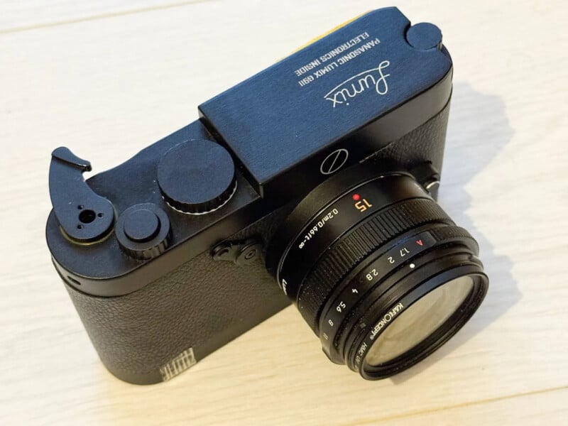 A black LTM-mount film camera labeled "Jumix" with a 15mm f/2.8 lens sits on a light wood surface, shown from an angled top-down perspective.