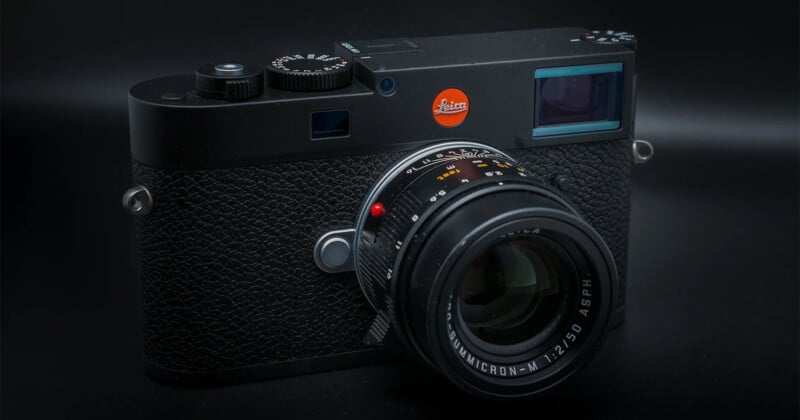 A close-up of a black Leica camera with a textured grip and a lens attached, set against a dark background. The red Leica logo is visible on the top right of the camera body.