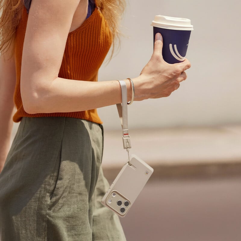 A person wearing an orange sleeveless top and green pants holds a navy blue disposable coffee cup in one hand and a white smartphone with a wrist strap in the other. The background is blurred.