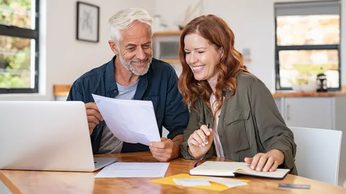 Mature couple sitting at a table, holding papers and working on their finances on a laptop in their house