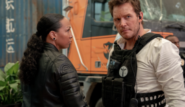A Black woman wearing a black leather jacket with her hair tied back in a long ponytail, eyes a white man suspiciously. He is wearing a white shirt with a black tactical vest on top.
