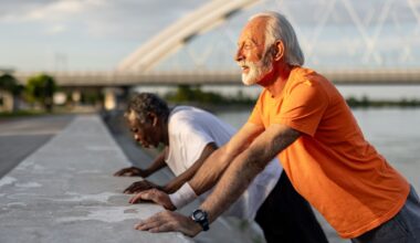 man in an orange tshirt and man in a white tshirt behind him both sideways to the camera performing press ups against a low wall outside.