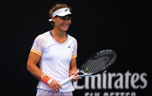 A Caucasian woman in her thirties appears in white tennis gear holding a tennis racket and wearing a visor. She is smiling