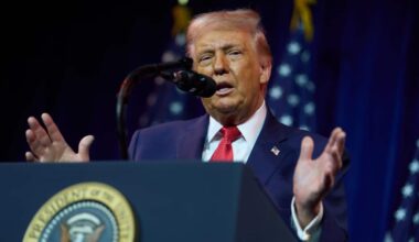 Donald Trump speaks from behind a presidential podium with his hands raised. Behind him are several US flags.