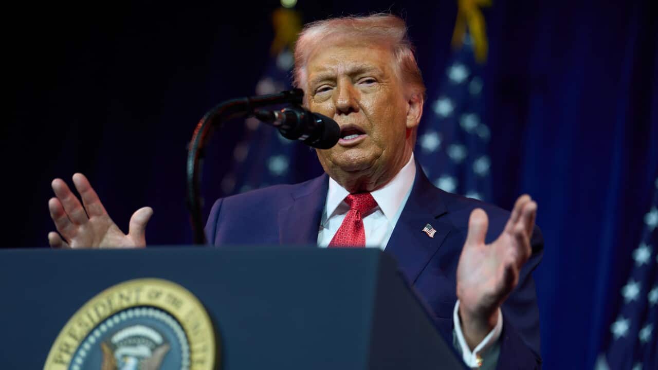 Donald Trump speaks from behind a presidential podium with his hands raised. Behind him are several US flags.