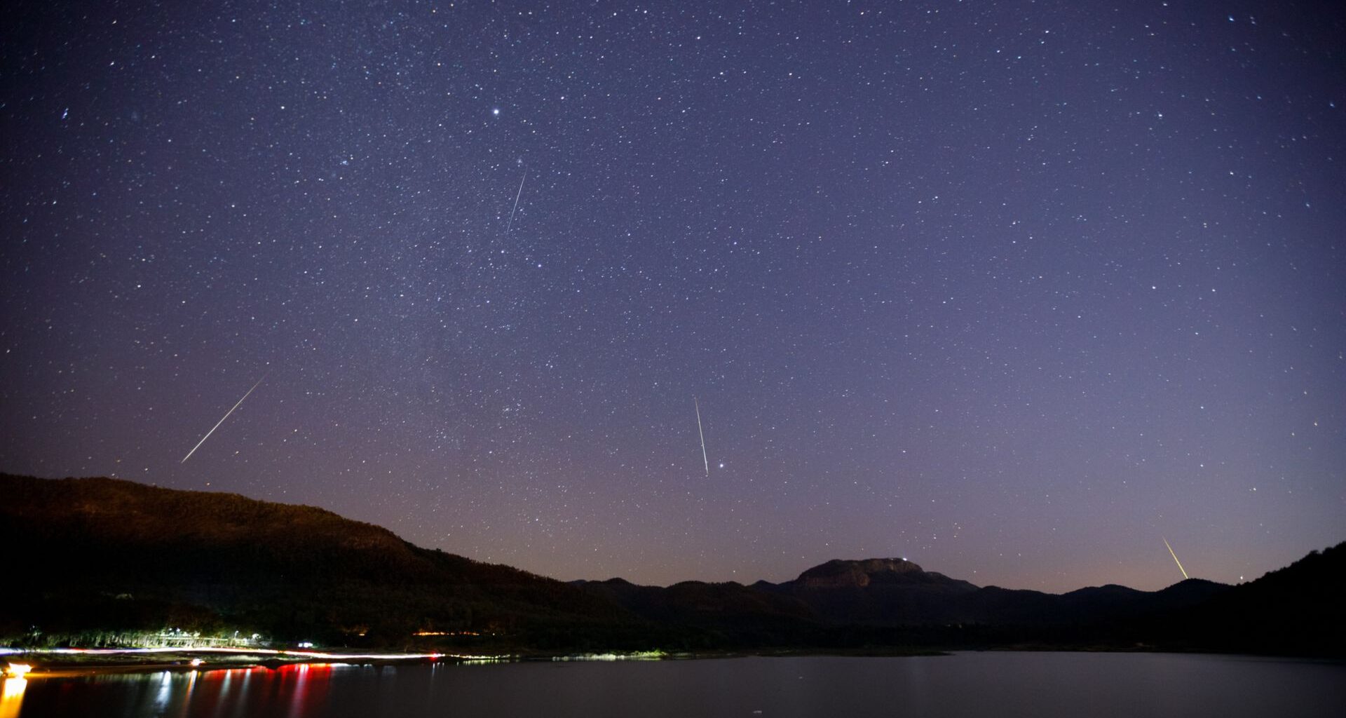 White streaks of meteors are seen against a dark purple night sky with a frozen lake with lights below pushed against silhouetted mountains in the background