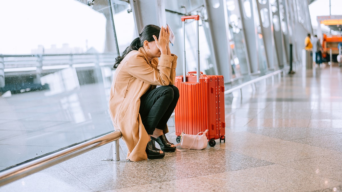 Woman sitting on the airport floor beside a suitcase, covering her face in distress.