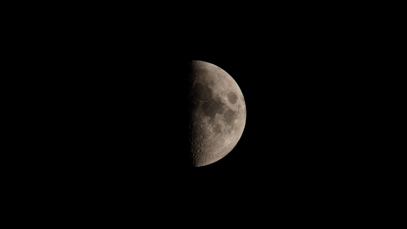 A photo of the moon captured during its first quarter phase it its right half sits illuminated by direct sunlight against a black night sky.