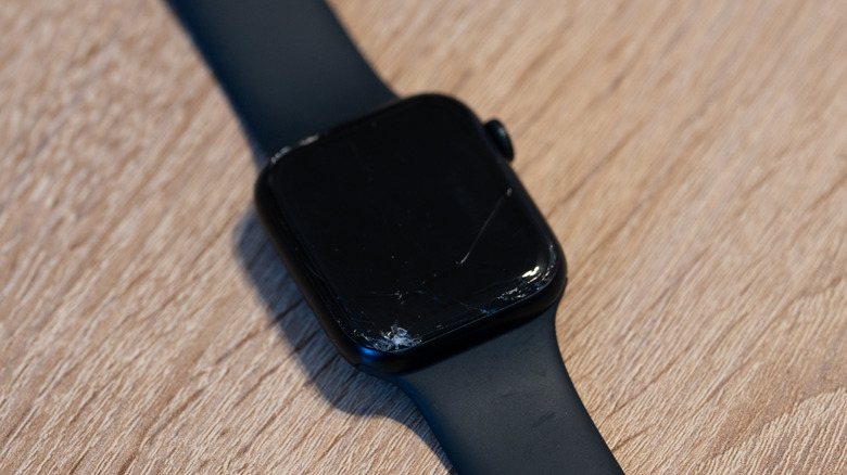 An Apple Watch with a black band and a cracked, damaged screen on a wood surface
