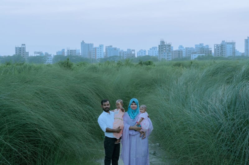 A family of four stands together in a field of tall green grass, with city buildings visible in the background under a cloudy sky. The adults hold two young children, and the woman wears a blue headscarf and purple dress.